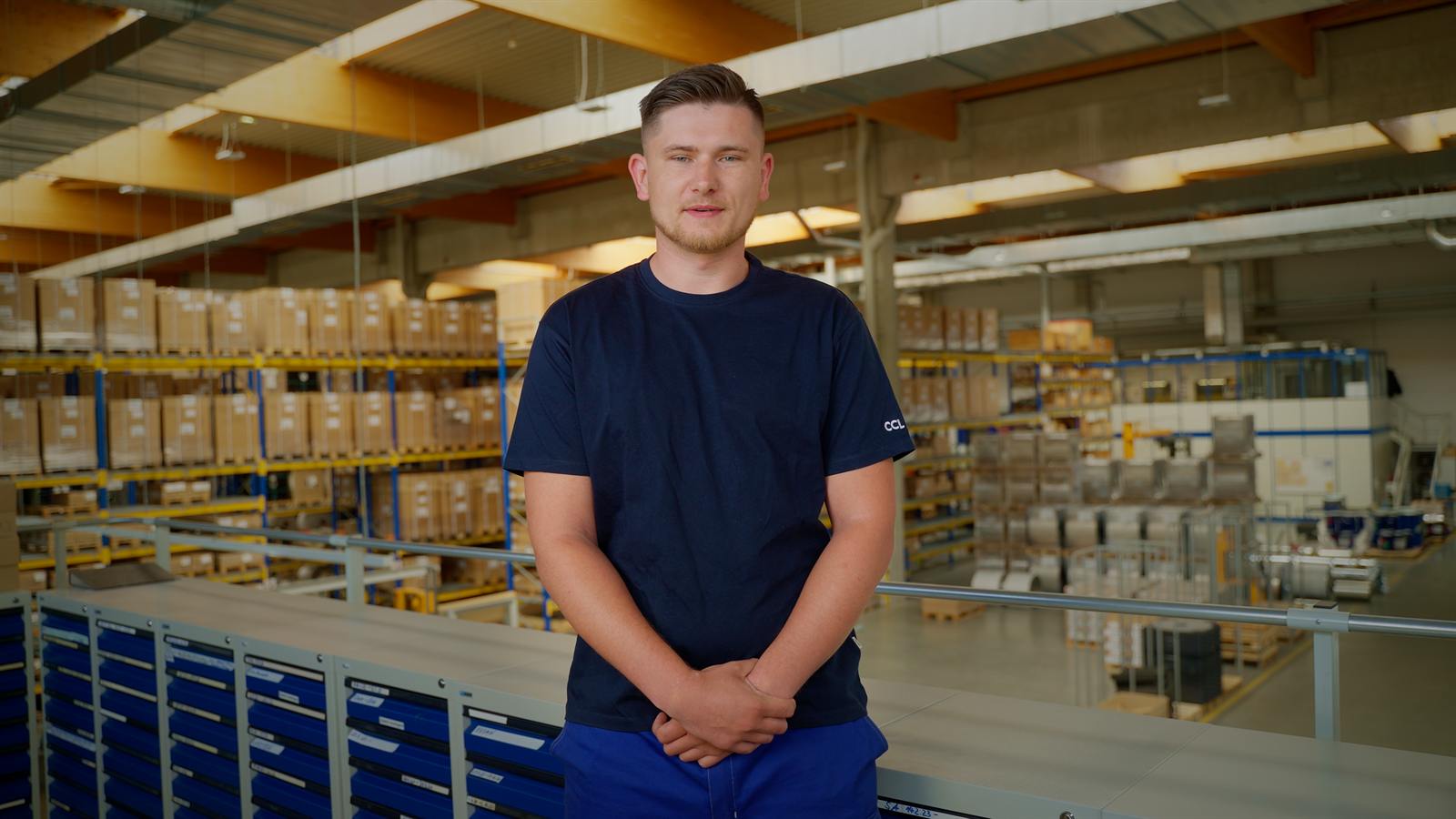 Mitarbeiter im blauen Shirt steht in der Produktionshalle und blickt in die Kamera